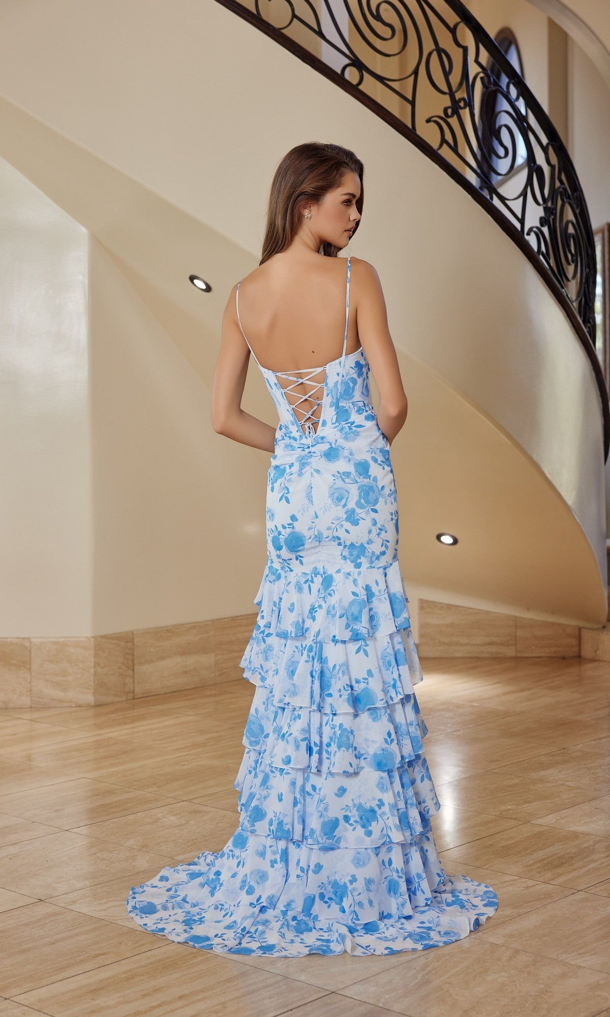 Woman in a long blue and white Nox Anabel T1863 prom dress with floral chiffon and lace-up back, standing indoors on a polished floor near a curved staircase.